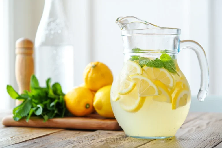 Limonade maison fraîche servie dans un pichet en verre avec des tranches de citron, des feuilles de menthe et des glaçons. Arrière-plan : citrons, menthe fraîche, bouteille d’eau pétillante et presse-citron en bois sur une table en bois clair. Recette estivale légère, sans alcool, faible en calories.