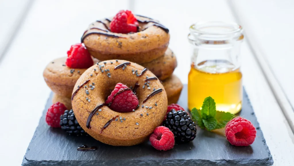 recette donuts moelleux et dorés cuits au four, décorés de glaçage coloré et parsemés de sprinkles. Servis sur une assiette blanche ou un plateau en bois, avec des touches créatives comme des copeaux de chocolat ou de la noix de coco râpée. Une texture légère et appétissante mise en valeur par une lumière douce.